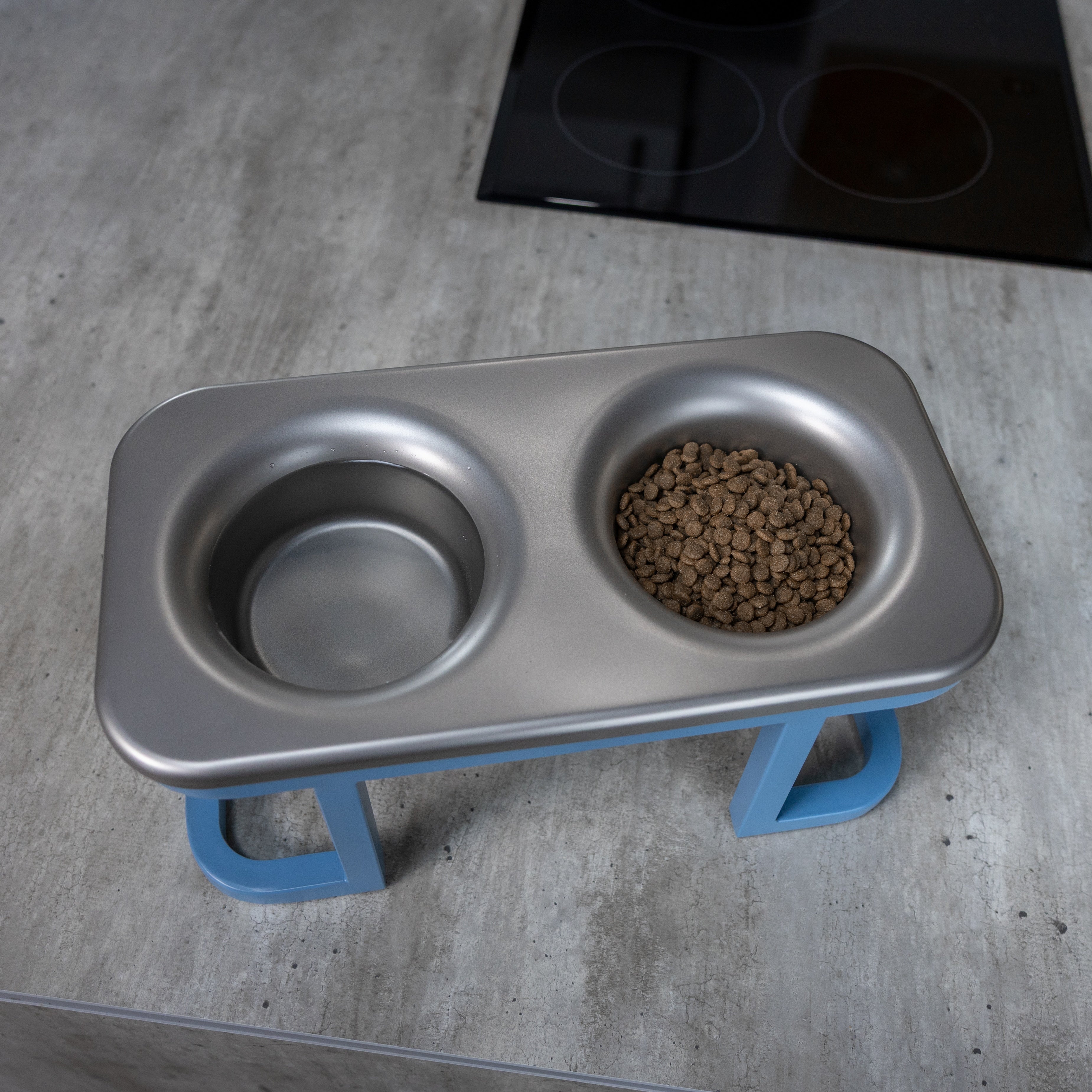 Metal double-bowl pet feeder with blue stand on a concrete countertop. No bacteria. No mess.